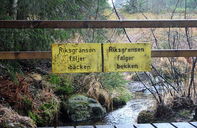 Bilde av en bro med to skilt, et skilt står det "Riksgrensen følger bekken" på norsk, det andre skiltet står det samme på svensk. 